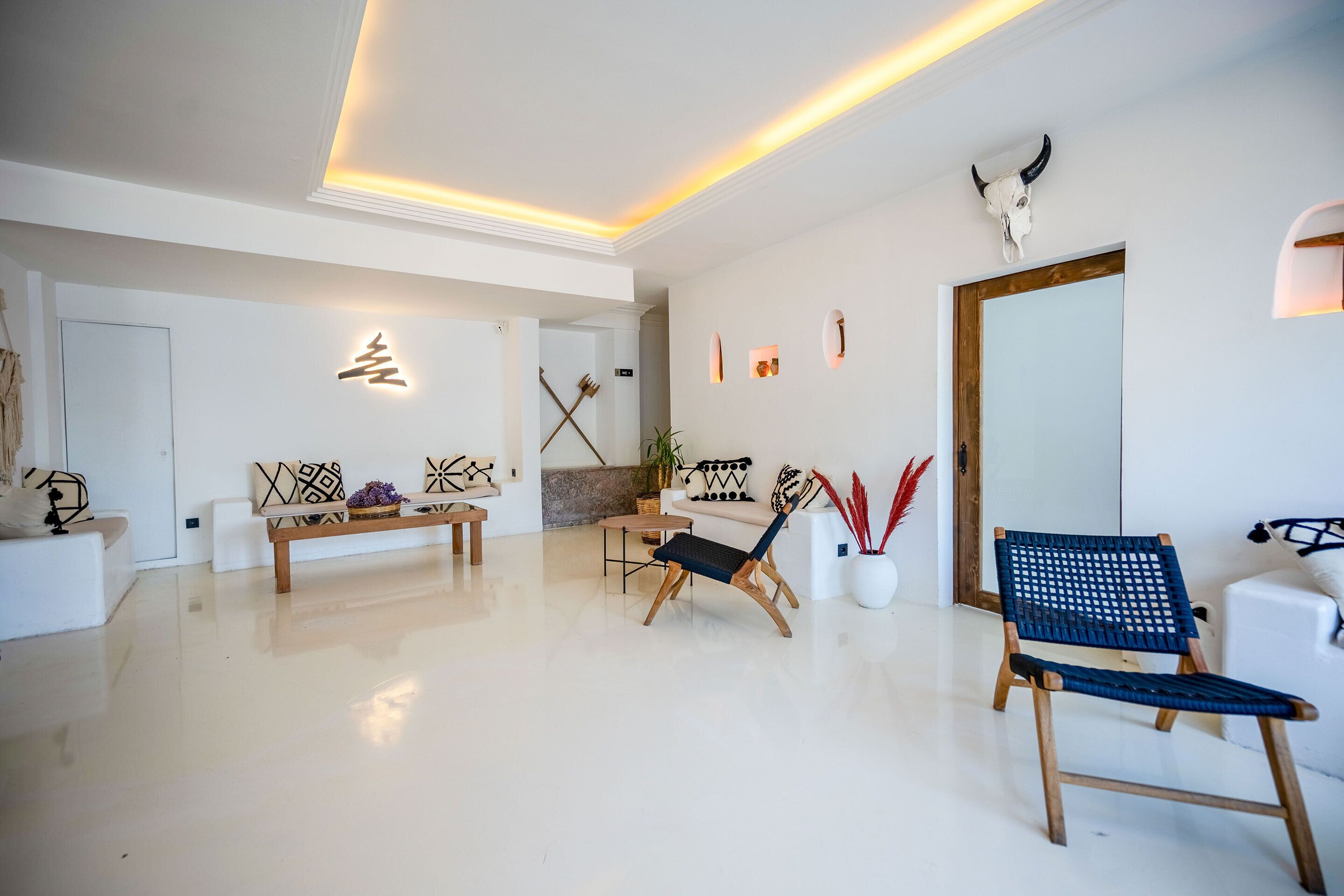 Una sala de estar moderna con un sofá blanco, un banco de madera y dos sillas. Hay un espejo grande en la pared y un cráneo decorativo en el techo.