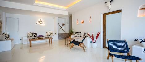 Una sala de estar moderna con un sofá blanco, un banco de madera y dos sillas. Hay un espejo grande en la pared y un cráneo decorativo en el techo.