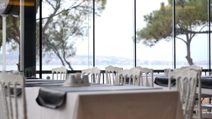 Un restaurante con sillas y mesas blancas, un letrero que dice "LILIK 87 HOTEL", y una vista de árboles y un paisaje urbano lejano a través de amplias ventanas.