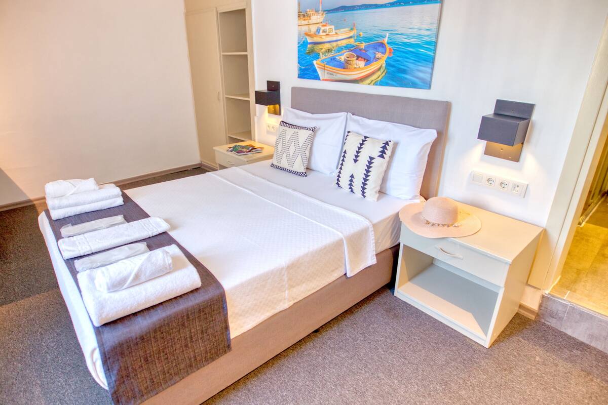 Un lit bien fait, avec du linge de lit blanc et un banc en bois, une table de chevet avec une lampe, un tableau représentant des bateaux accroché au mur et une petite étagère sur laquelle repose un chapeau.