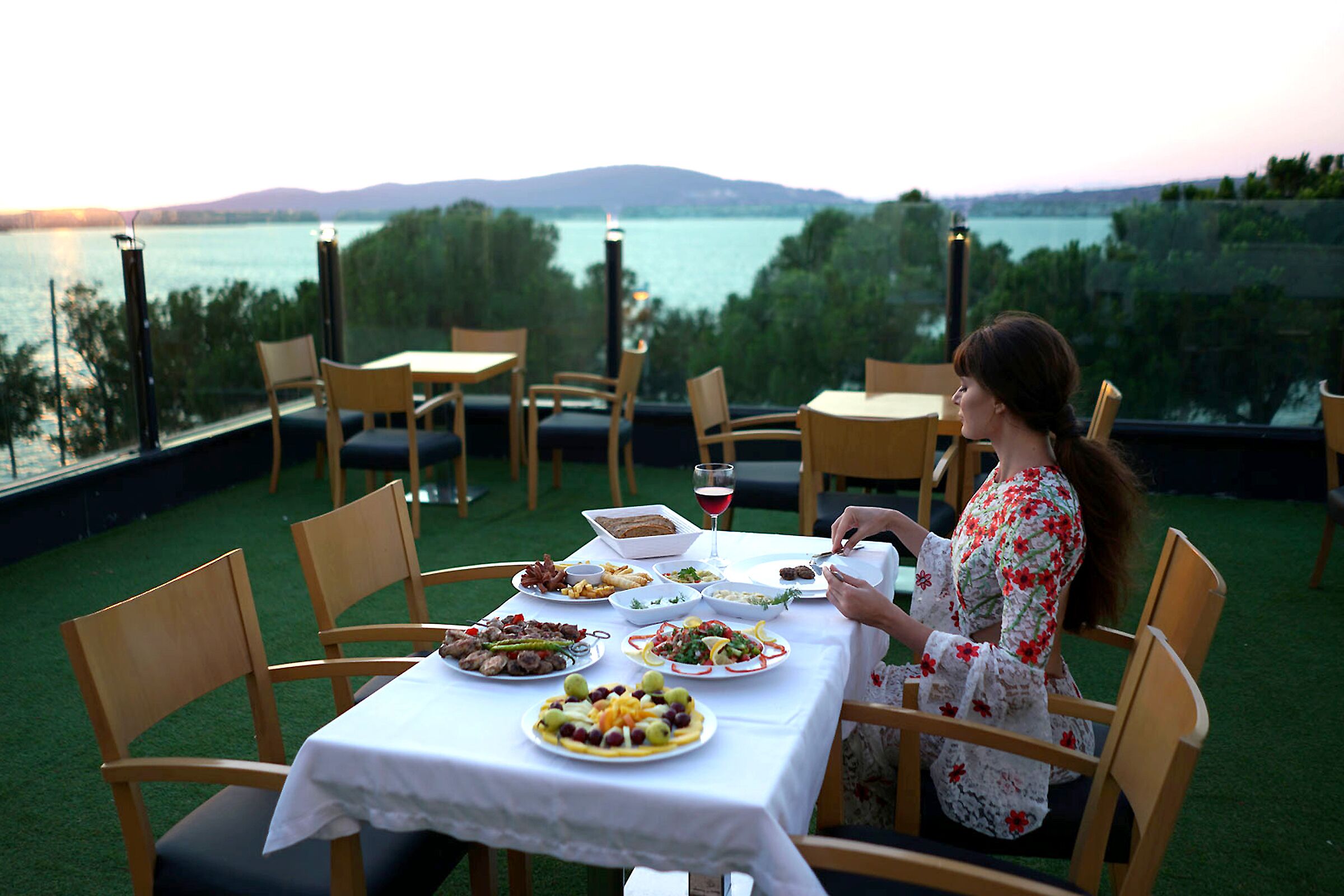 An outdoor dining area with a table set for a meal, surrounded by chairs, and overlooking a body of water with mountains in the distance.