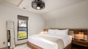 A modern bedroom with a large bed, a wooden headboard, a bedside table with a lamp, a window with blinds, and a white brick wall. - *Downtown Luxury + Outdoor Patio*~ Walk Everywhere (Bozeman)
