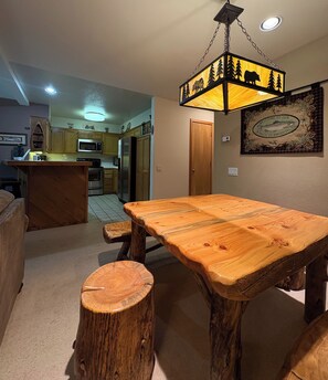 Un comedor con una mesa de madera y bancos, una lámpara colgante y una cocina al fondo.