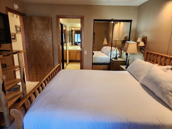 A hotel room with a large bed, wooden headboard, a wooden wardrobe, a mirror, a bedside table with lamps, and a view into the bathroom.
