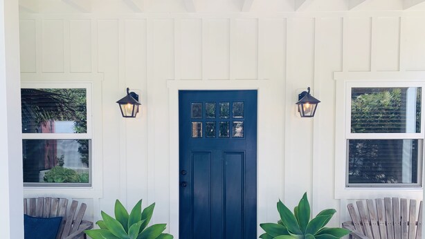 A front porch with a dark blue door, two potted plants, two wooden chairs, and two wall-mounted lanterns. - Blue Door Bungalow.  (Montecito)