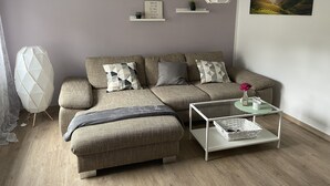 A modern living room with a sofa, ottoman, and a coffee table. There are decorative pillows, a wall-mounted shelf with a plant, and a hanging light fixture. - Ferienwohnung mit Balkon (Bruttig-Fankel)