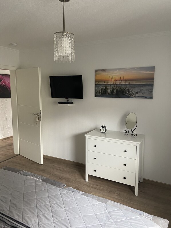 A bedroom with a bed, a TV, a dresser, a mirror, and a painting of a sunset.