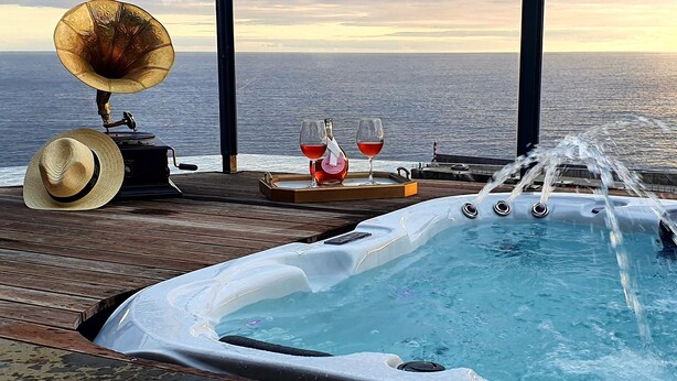 Un bain à remous avec jets d’eau, deux verres de vin rouge, un gramophone et un chapeau de paille sur une terrasse en bois offrant une vue sur l’océan.