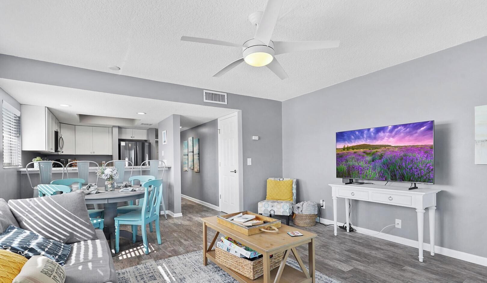 A modern living room with a dining area, a ceiling fan, a flat-screen TV, and a kitchen in the background.