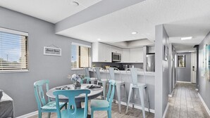 A modern kitchen with white cabinets, a microwave, and a refrigerator. There is a dining area with a round table and blue chairs. - 2 Bedroom Pet-friendly ocean view condo - OV3 (St Augustine)