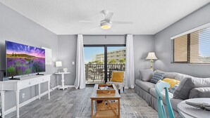 A modern living room with a flat-screen TV, a ceiling fan, a balcony with a view of a building, and a sofa with cushions. - 2 Bedroom Pet-friendly ocean view condo - OV3 (St Augustine)