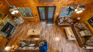 Interior de una cabaña con chimenea de piedra, suelo de madera, dos sofás, una mesa de centro y un cartel que dice 'WELCOME TO THE CABIN'.