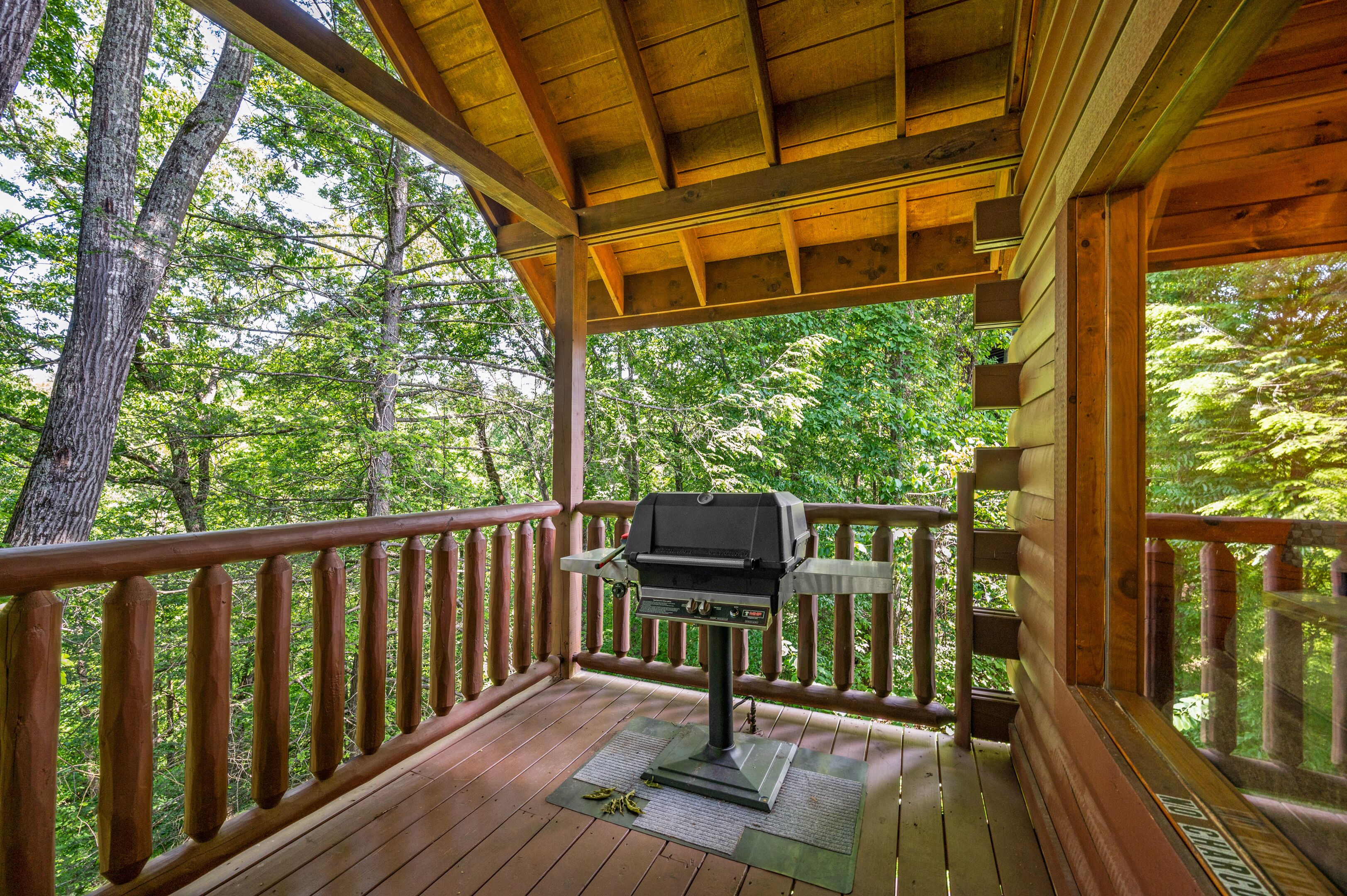 A wooden deck with a barbecue grill, surrounded by a wooden railing and trees.