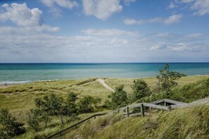 Et kystlandskab med en græssti, der fører hen til en træbygning, med udsigt over en strand og havet.