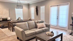 A modern living room with a leather sofa, a coffee table, a dining table, and a chandelier. - Seasonal Beach Service & Heated Pool + Golf Cart Included!! (Destin)