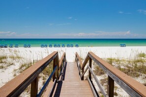 A wooden boardwalk leading to a beach with blue umbrellas and a clear blue sky.