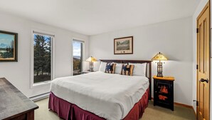 A bedroom with a large bed, two bedside lamps, a wooden nightstand, a framed picture on the wall, and a window with a view of trees.