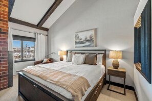 A bedroom with a large bed, two bedside tables with lamps, a window with curtains, a framed picture on the wall, and a view of a snowy landscape. - Bear Claw 510 Exceptional Mountain Condo with Ski-In/Ski-Out Access! (Steamboat Springs)