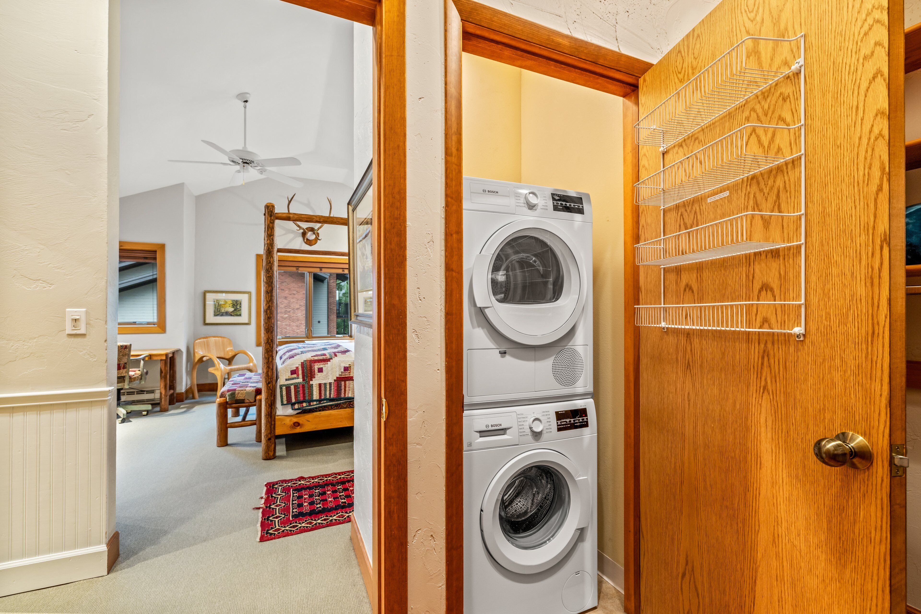 A laundry room with a Bosch washer and dryer, a wooden door with a metal rack, and a view into a bedroom with a bed, a chair, and a rug.