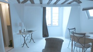Et værelse med et rundt bord, en stol, et lille bord med en lampe og et vindue med gardin.