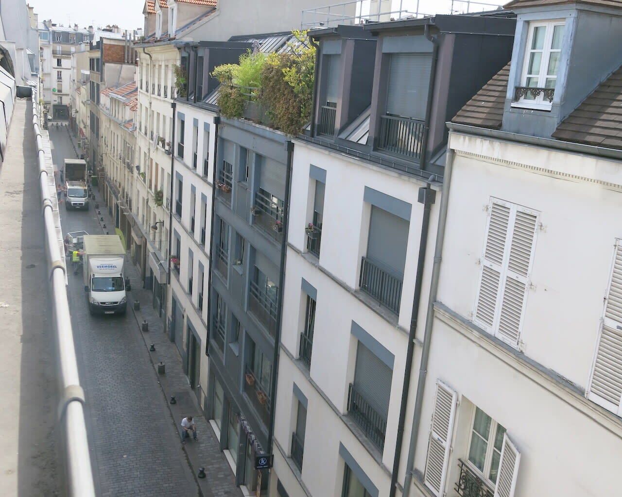 A narrow urban street lined with multi-story buildings, some with balconies and shutters. A white van is parked on the street.