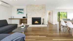 A modern living room with a fireplace, wooden flooring, a sofa, a dining area with chairs, and a view of greenery outside. - Vue Sur La Mer (Wye River)