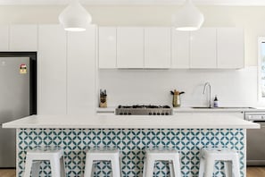 A modern kitchen with white cabinets, a central island with bar stools, a gas stove, and pendant lighting. - Vue Sur La Mer (Wye River)