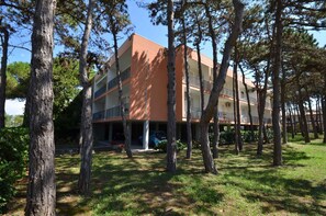 A multi-story building with a flat roof and balconies, surrounded by trees and a grassy area.