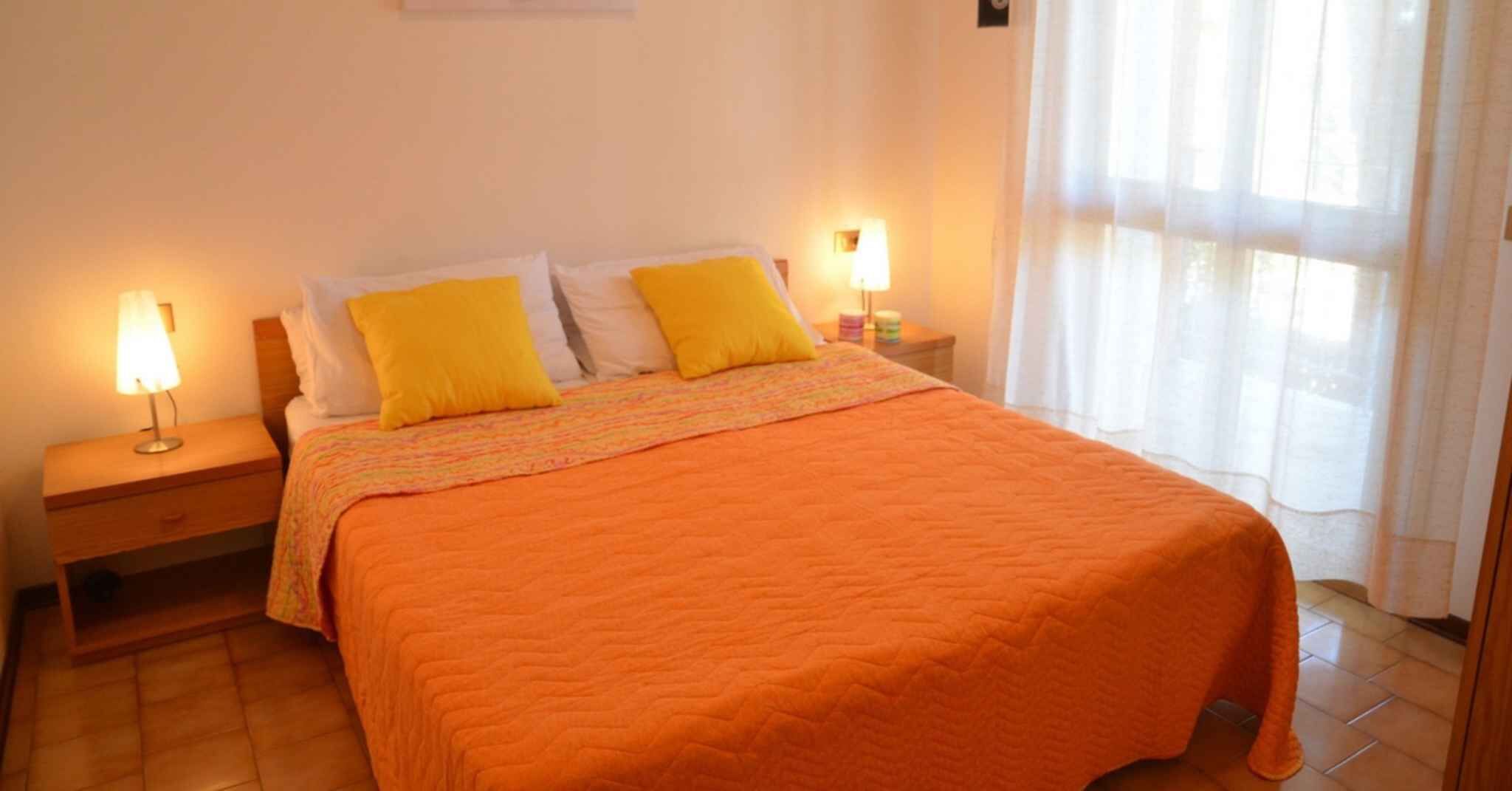 A neatly made bed with an orange bedspread and two yellow pillows, flanked by nightstands with lamps, in a room with a window and curtains.