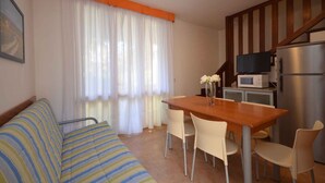 Ein kompakter Wohnbereich mit Sofa, Esstisch, Stühlen, einer Küchenzeile inklusive Mikrowelle und Kühlschrank sowie einer Treppe.