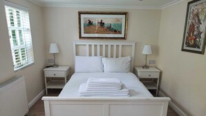 Un dormitorio con una cama blanca, dos mesitas de noche, un radiador y una ventana con persianas.