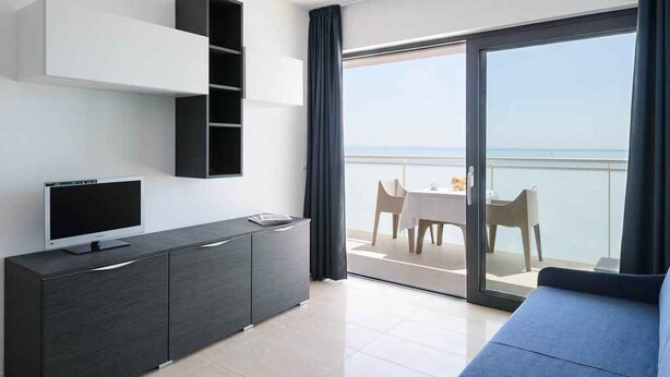 Una habitación de hotel moderna con balcón, televisor de pantalla plana, sofá de color oscuro y zona de comedor con mesa y sillas.