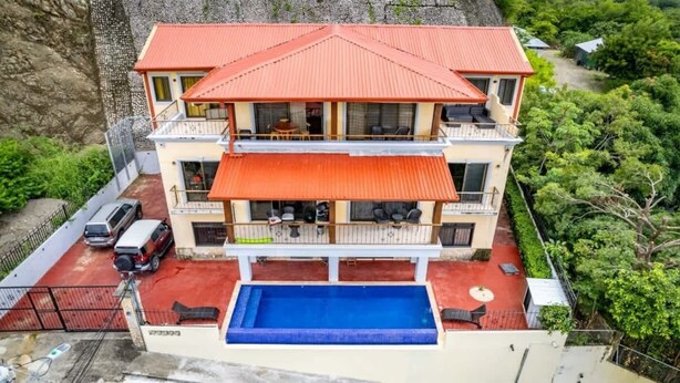 Un edificio de dos pisos con techo rojo, piscina y balcón con mobiliario exterior.