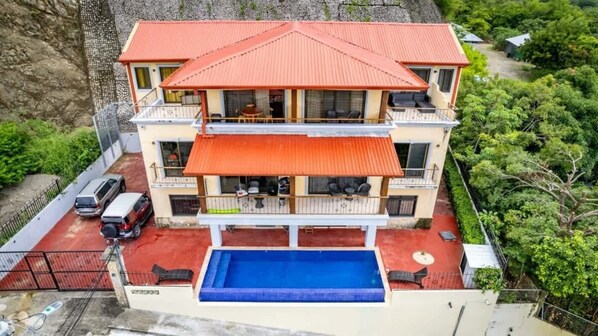 A two-story building with a red roof, a swimming pool, and a balcony with outdoor seating.