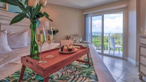 A spacious room with a large window, a wooden dining table with a tray of pastries, and a vase with flowers. - Dazzling Ocean View Suite w/heated pool, beach, marina, tennis/pickleball (Tavernier)