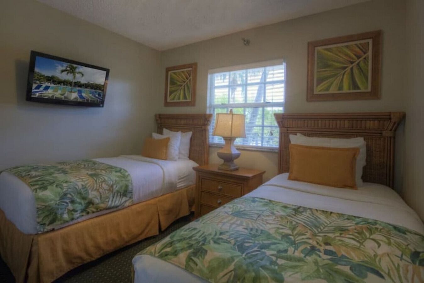 Een hotelkamer met twee bedden, een nachtkastje met lamp, een raam met luxaflex en een schilderij van een tropisch landschap.