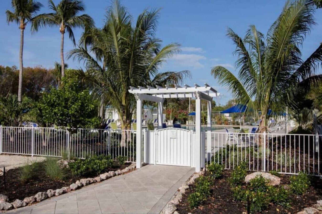 A gated entrance with a white pergola, palm trees, and a pool area.
