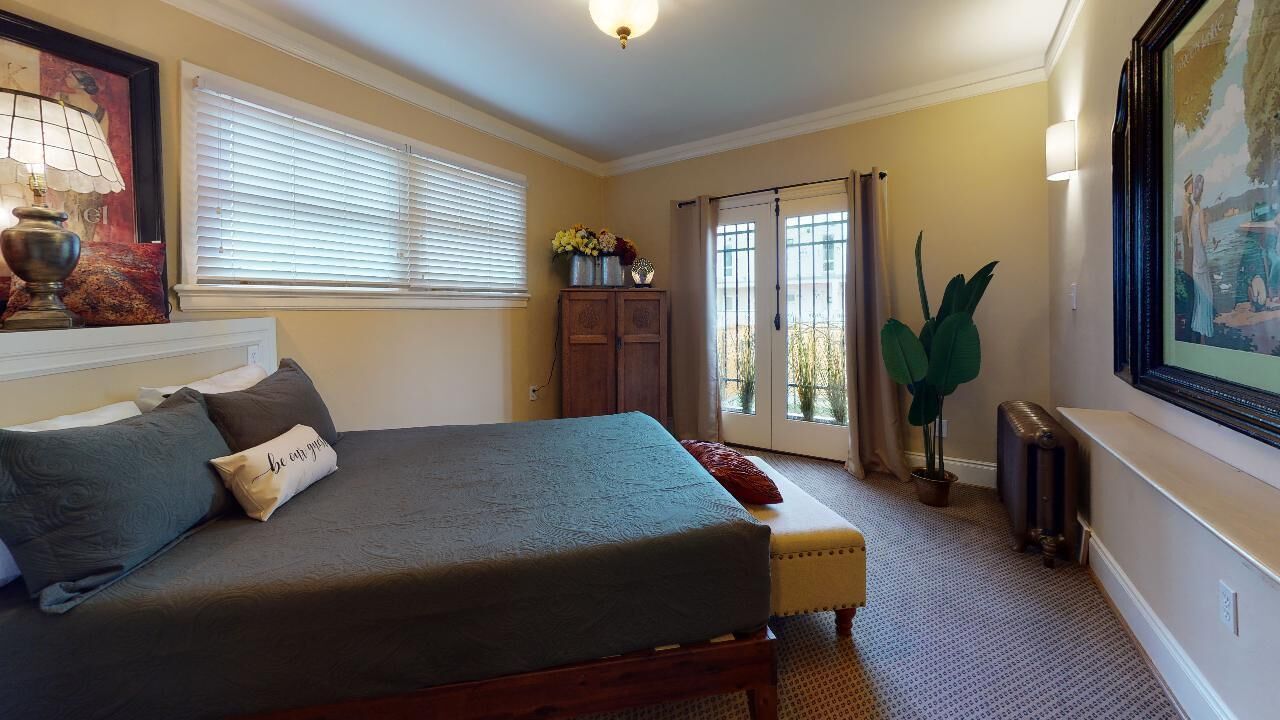 A bedroom with a bed, a nightstand, a dresser, a window with blinds, a radiator, and a potted plant.
