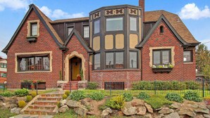 A large, two-story brick house with a prominent bay window, a stone staircase leading to the front door, and a well-maintained garden with a variety of plants and a small pond. - Lower Suite: a classic suite in a quiet Seattle home (Seattle)