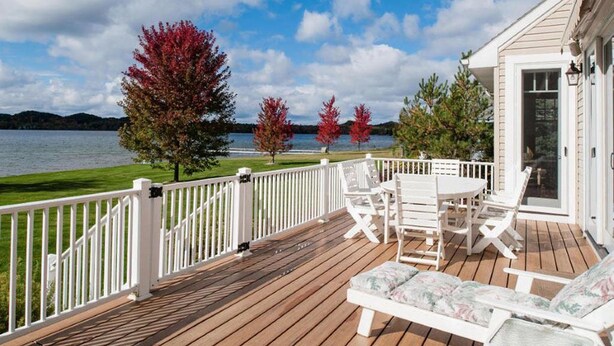 A wooden deck with white railings, a table with chairs, and a view of a lake with autumn trees.