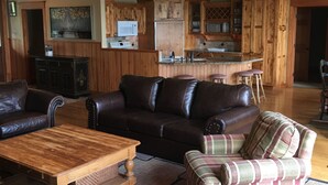 A spacious living room with a dark leather sofa, a wooden coffee table, and a plaid armchair. A kitchen with wooden cabinets and a bar area is visible in the background.
