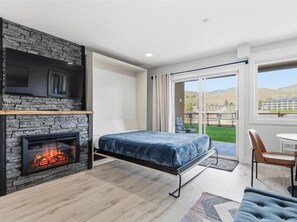 A bedroom with a stone fireplace, a bed, a chair, and a view of a grassy area through a sliding glass door.