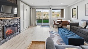 Een moderne woonkamer met een stenen open haard, een bank, een televisie en uitzicht op een groen landschap door grote ramen.
