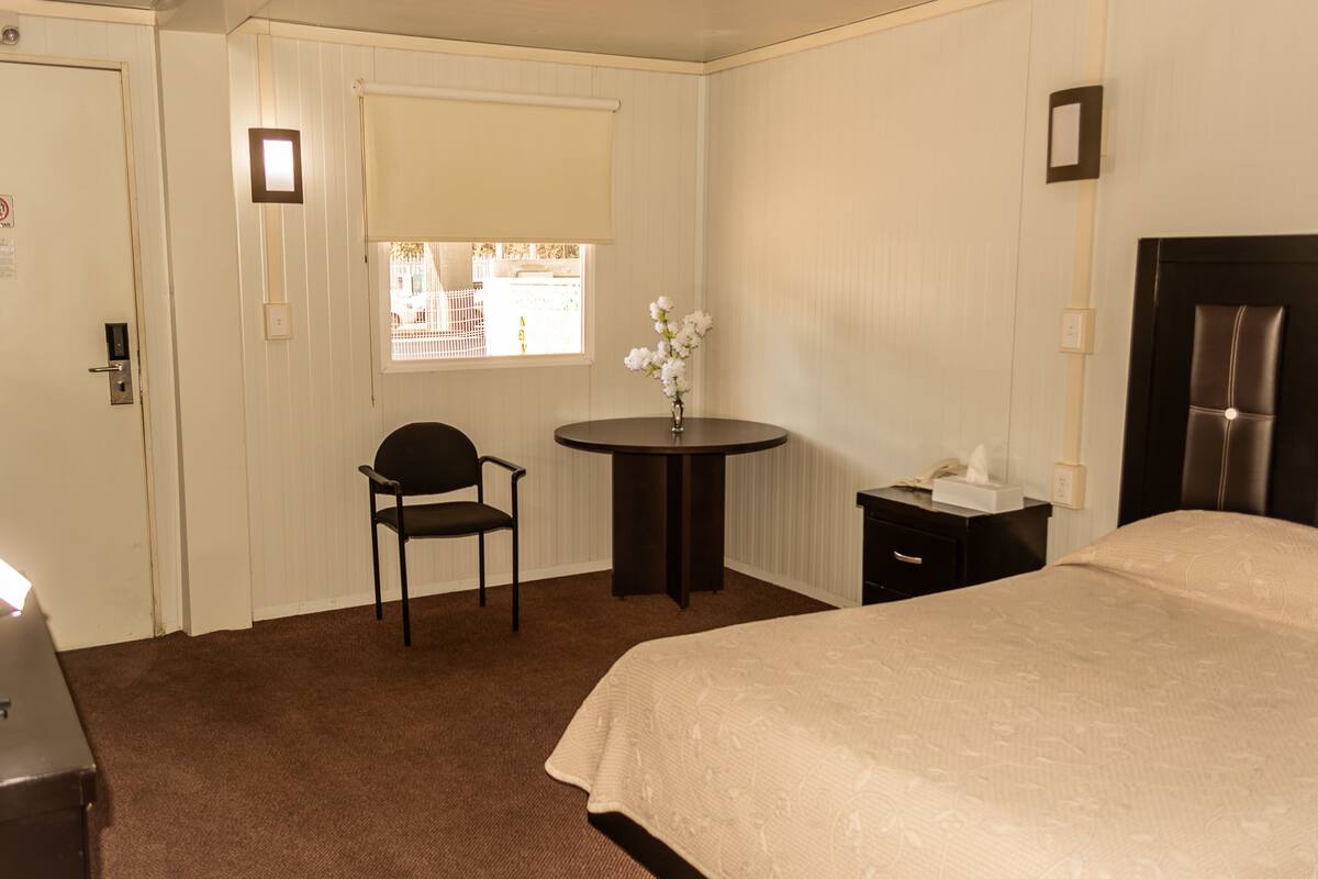 A hotel room with a bed, a chair, a small table with a vase, and a window.