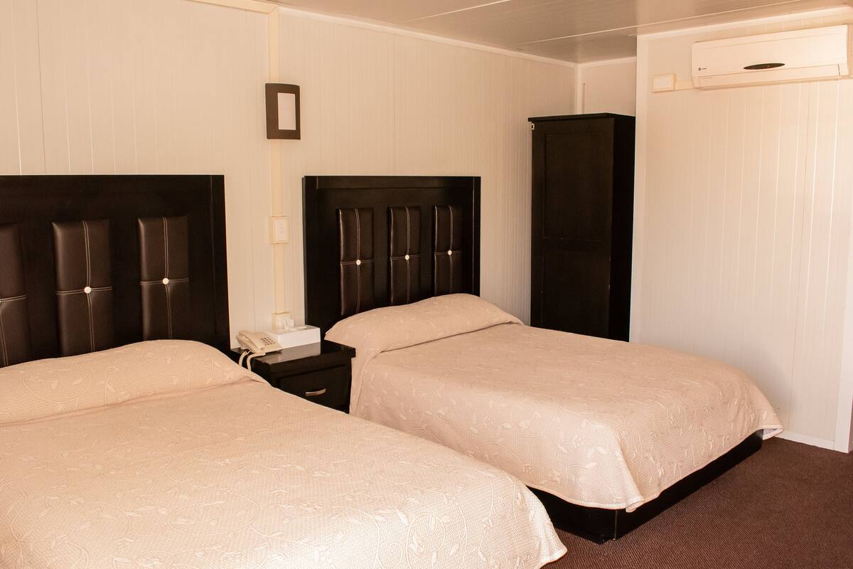 A hotel room with two beds, a wardrobe, a bedside table with a phone, and an air conditioning unit.