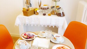 Una mesa servida con alimentos para el desayuno, como cruasanes, pasteles y café, con mantel blanco y sillas color naranja.