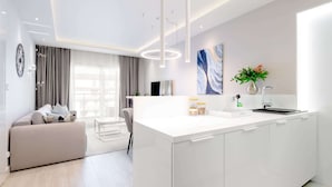 Una cocina moderna con cubierta blanca, horno empotrado y un lavamanos. Hay una zona de comedor con un sofá y una silla.