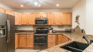 Una cocina con armarios de madera, un refrigerador de acero inoxidable, un microondas, un horno, encimeras de granito y un lavamanos negro.