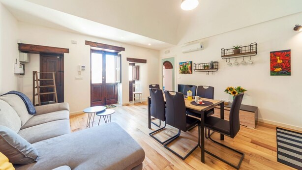 Uma sala de estar moderna com um sofá cinza, uma mesa de jantar com cadeiras e uma cozinha visível por uma porta aberta.