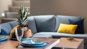 Un comedor con una mesa de madera dispuesta para dos personas, con platos, una copa y una piña. En el fondo, un sofá con cojines.